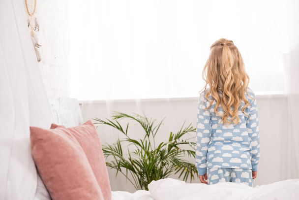 back view of child in pajamas standing near window in bedroom - Foto, imagen