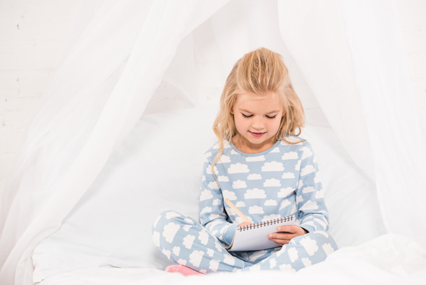 αξιολάτρευτο παιδί στο πιτζάμες κάθεται στο κρεβάτι με σταυρωμένα πόδια και γράφοντας στο Σημειωματάριο - Φωτογραφία, εικόνα
