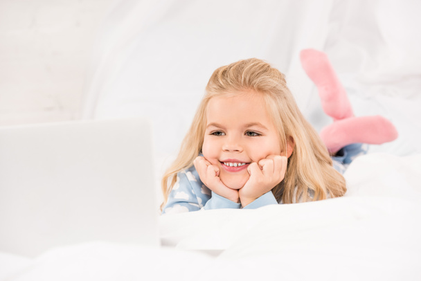 niño adorable feliz acostado en la cama y viendo dibujos animados en el ordenador portátil
 - Foto, imagen