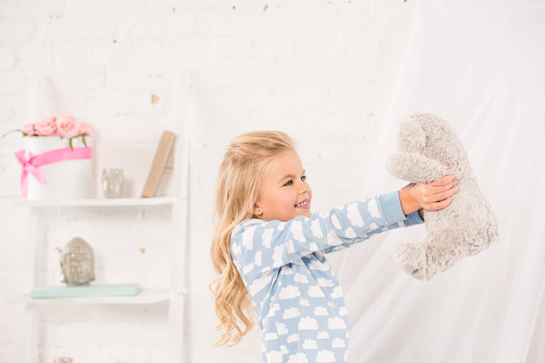 Lächelndes süßes Kind spielt mit Teddybär im Schlafzimmer - Foto, Bild