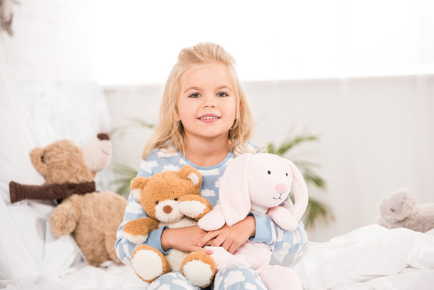 усміхнена чарівна дитина сидить з м'якими іграшками на ліжку
 - Фото, зображення