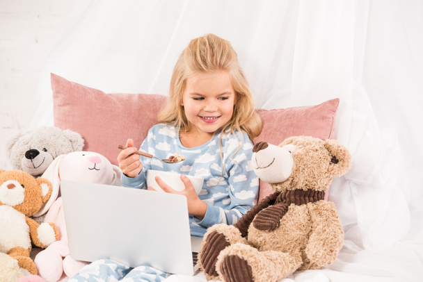 cute child with laptop eating cornflakes and looking at teddy bear in bed - Фото, зображення