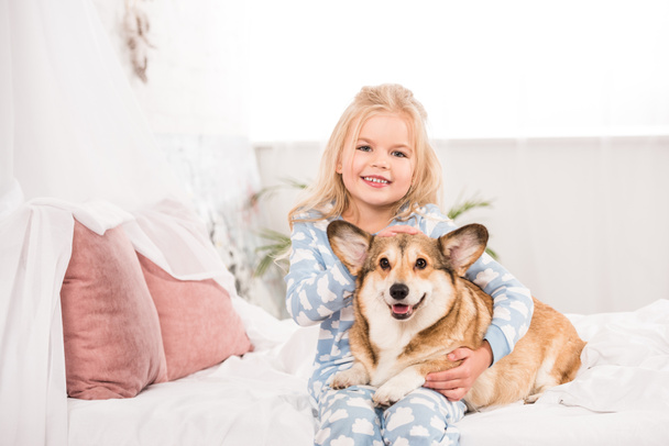 smiling child in pajamas embracing and petting corgi dog in bed - Fotografie, Obrázek