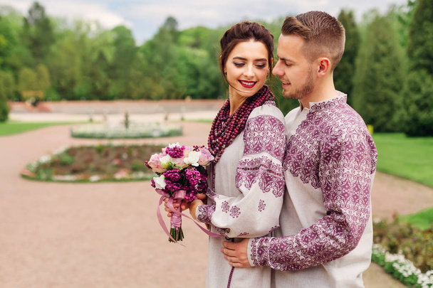 boldog ifjú házasok hímzett ruhát mosoly, a háttérben a Park. a menyasszony a csokrot tartja, és a vőlegény hugs neki a hátsó oldalán - Fotó, kép