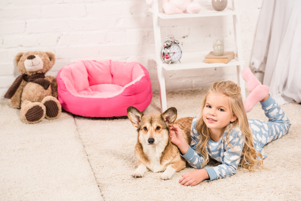 Lächelndes Kind im Schlafanzug blickt in die Kamera und streichelt den welschen Corgi-Hund zu Hause  - Foto, Bild