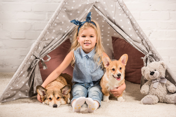 cute child looking at camera and hugging welsh corgi dogs in wigwam at home - Fotografie, Obrázek