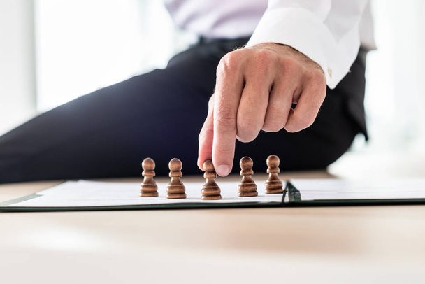 Líder de negocios sentado en su escritorio tomando decisiones de recursos humanos colocando piezas de ajedrez de peón en el papeleo
. - Foto, Imagen
