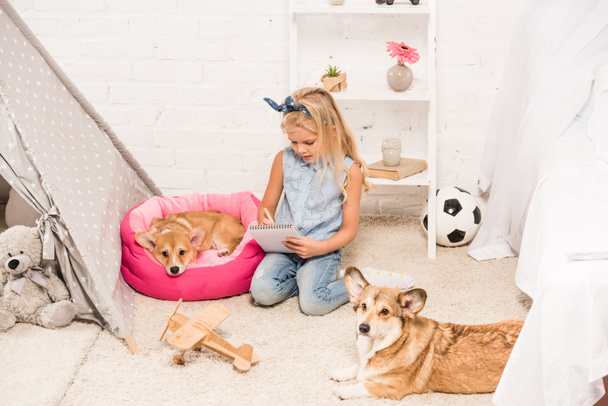 cute child sitting at home with welsh corgi dogs and writing in notebook - Fotó, kép