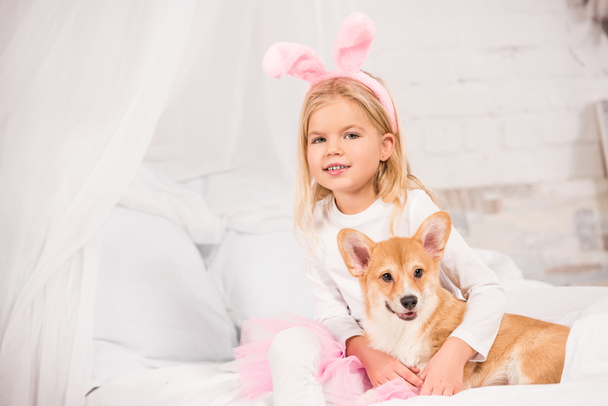 uśmiechnięte dziecko w bunny uszy pałąk siedzi z welsh corgi psa na łóżko w domu  - Zdjęcie, obraz