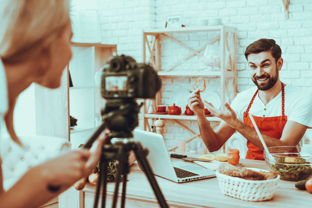 Blogger Makes a Video. Blogger is Smiling Beard Man. Video About a Cooking. Woman Operator Shoots a Video on Camera. Different Food on Table. Man Showing a Piece of Pepper. People in Studio Interior. - Photo, Image