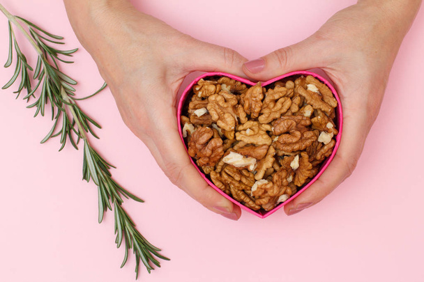 Woman holding a box in heart shape with peeled walnuts on pink background with rosemary. Celebration day concept. Useful nutritious protein product. Top view. - Photo, Image