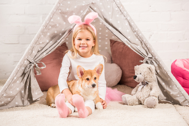 милый ребенок в кроличьих ушах повязка сидя с корги собака и плюшевый мишка в wigwam
 - Фото, изображение