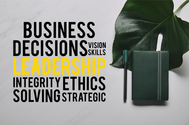 grünes Notizbuch mit Stift auf weißem Marmorhintergrund mit Monstera-Blatt und unternehmerischen Entscheidungen, Visionen und Führungsqualitäten - Foto, Bild