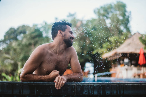 Portrait of a young handsome man in an outdoor pool in the tropics. Attractive guy with a beard at the side of the pool - Foto, Bild