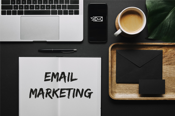 Ανοίξτε το σημειωματάριο με την επιγραφή «μάρκετινγκ ηλεκτρονικού ταχυδρομείου» από χαρτικά και lap-top σε μαύρο φόντο - Φωτογραφία, εικόνα