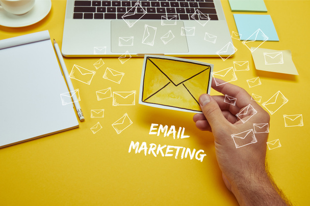 cropped image of man holding envelope sign near laptop with "email marketing" lettering and email icons - Foto, imagen