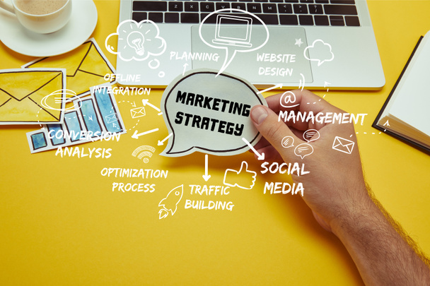imagem cortada do homem segurando bolha de fala com lettering "estratégia de marketing" e ícones perto do laptop
 - Foto, Imagem