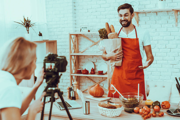 Happy Man Blogger Makes a Video. Video About a Cooking. Woman Operator Shoots a Video on Camera. Different Food on Table. Man Holding a Shopping Bag with Products. People in Studio Interior. - Zdjęcie, obraz