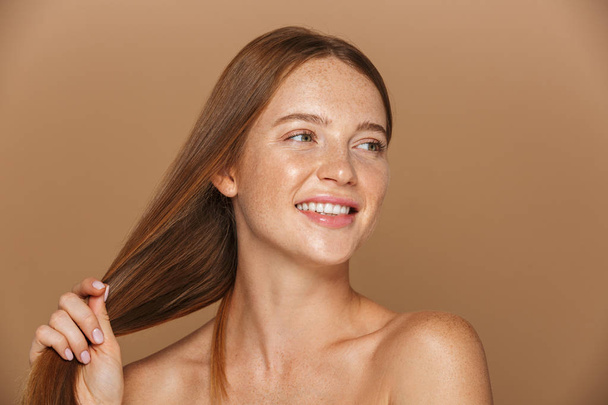 Beauty portrait of a smiling young topless woman with long red hair posing, playing with her hair isolated over beige background - Fotoğraf, Görsel