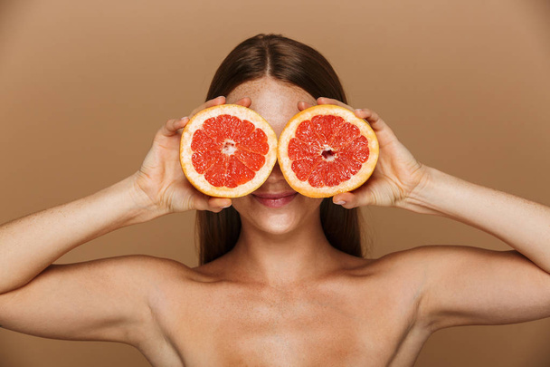 Beauty portrait of a beautiful healthy young topless woman with long red hair showing sliced grapefruit isolated over beige background - Foto, immagini