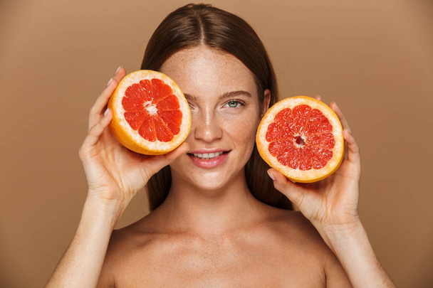 Beauty portrait of a beautiful healthy young topless woman with long red hair showing sliced grapefruit isolated over beige background - Φωτογραφία, εικόνα