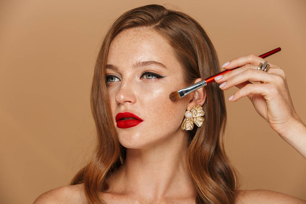 Close up of a beautiful young topless woman wearing makeup and jewelry accessories posing isolated over beige background, holding makeup brush at her face - Foto, immagini