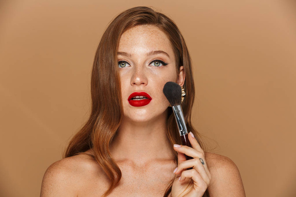 Close up of a beautiful young topless woman wearing makeup and jewelry accessories posing isolated over beige background, holding makeup brush at her face - 写真・画像