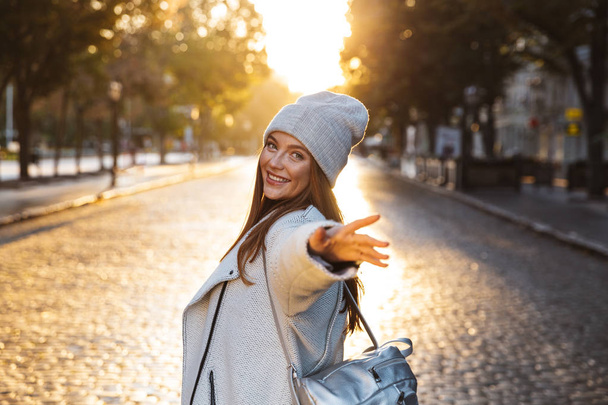 Cheerful young woman dressed in autumn coat and hat walking outdoors at the city street - Fotó, kép