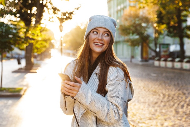 Beautiful young woman dressed in autumn coat and hat walking outdoors, using mobile phone - Foto, afbeelding