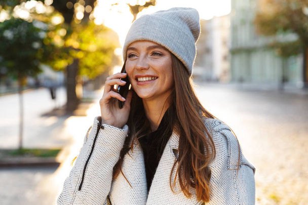 Image of a cheerful beautiful redhead woman walking outdoors wearing hat talking by mobile phone. - Fotó, kép