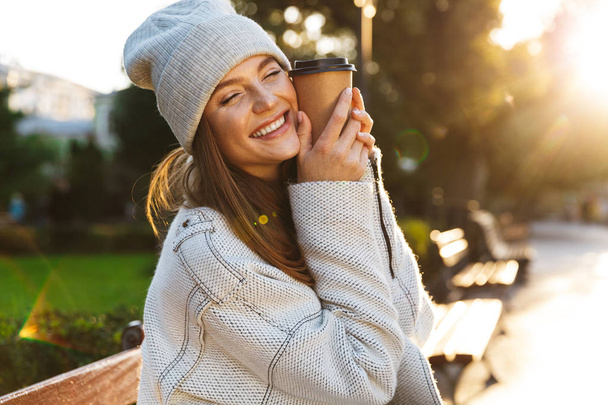 Beautiful young woman dressed in autumn coat and hat sitting on a bench outdoors, holding cup of warm coffee - Photo, image
