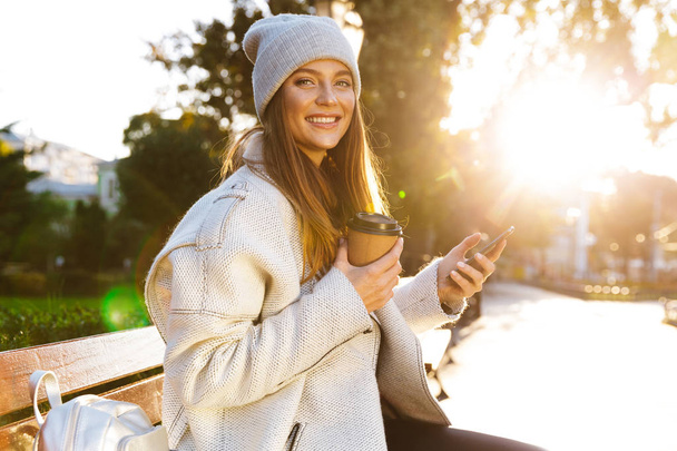 Beautiful young woman dressed in autumn coat and hat sitting on a bench outdoors, drinking coffee, using mobile phone - Foto, imagen