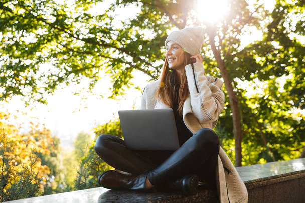 Beautiful young woman dressed in autumn coat and hat sitting outdoors, using laptop computer, celebrating success - Fotografie, Obrázek
