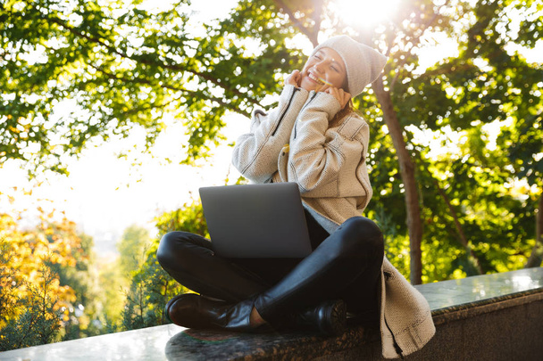 Beautiful young woman dressed in autumn coat and hat sitting outdoors, using laptop computer, celebrating success - Fotoğraf, Görsel