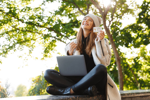 Happy young woman dressed in autumn coat and hat sitting outdoors, using laptop computer, showing plastic credit card - Φωτογραφία, εικόνα