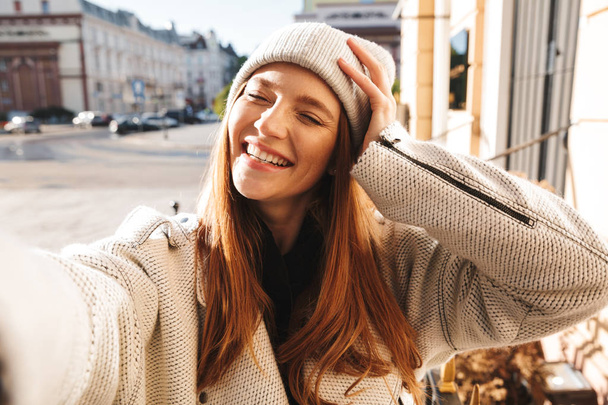 Beautiful young woman dressed in autumn coat and hat walking outdoors, taking a selfie - Foto, Imagen
