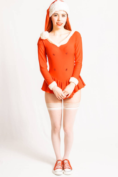 Young and pretty red head girl in a Santa Claus mini dress on a neutral white background. - Photo, Image