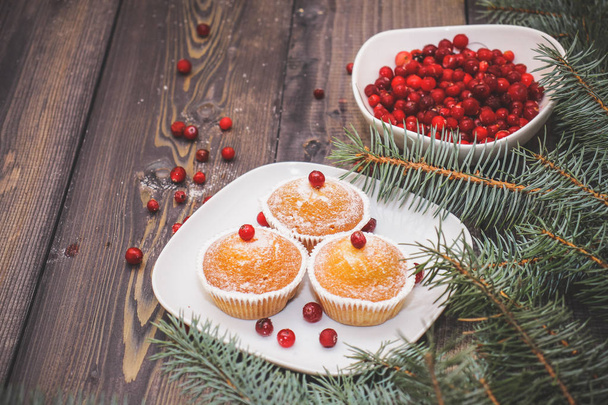 Festive composition for the background of New Year's postcards. A light wooden table top with a plate of freshly baked muffins decorated with red berries sprinkled with white powder and a plate of red berries with spruce twigs. - Photo, Image