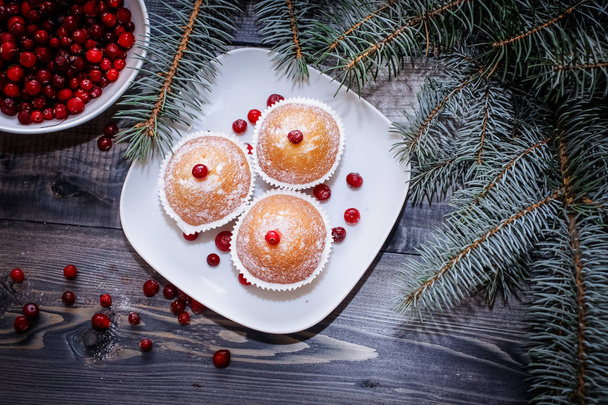 Festive composition for the background of New Year's postcards. A light wooden table top with a plate of freshly baked muffins decorated with red berries sprinkled with white powder and a plate of red berries with spruce twigs. - 写真・画像
