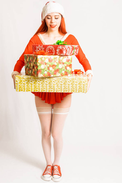 Chica joven y bonita cabeza roja en un mini vestido de Santa Claus sobre un fondo blanco neutro con regalos
 - Foto, Imagen