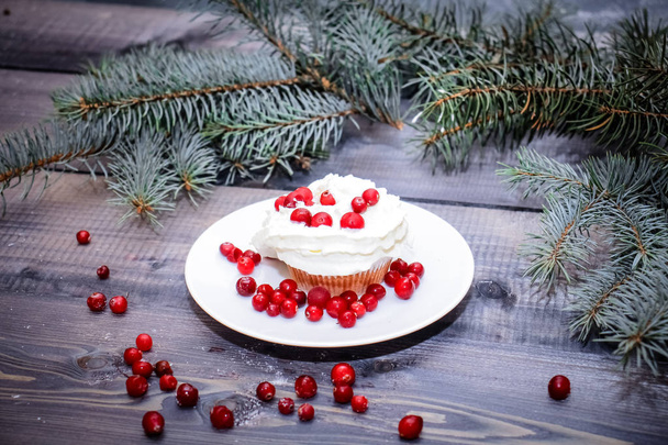 Festive composition for the background of New Year's postcards. Freshly baked cake on a white plate decorated with red berries and whipped white cream with a plate topped with red berries decorated with spruce sprigs on a light wooden table top. - Fotó, kép