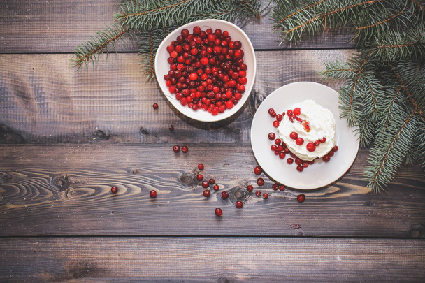 Festive composition for the background of New Year's postcards. Freshly baked cake on a white plate decorated with red berries and whipped white cream with a plate topped with red berries decorated with spruce sprigs on a light wooden table top. - Valokuva, kuva