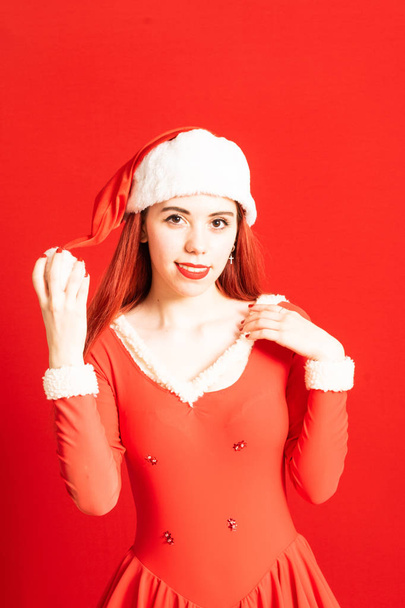 Chica joven y bonita cabeza roja en un mini vestido de Santa Claus sobre un fondo rojo neutro
. - Foto, Imagen