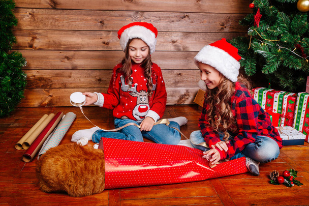fröhliche lustige kleine Schwestern in Weihnachtsmützen packen Geschenke in der Nähe des Weihnachtsbaums - Foto, Bild