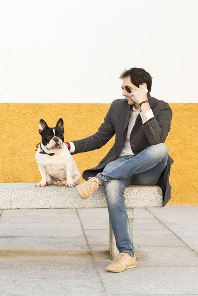 Urban scene of a man with a dog french bulldog sited on stone bench talking by phone. - Photo, image