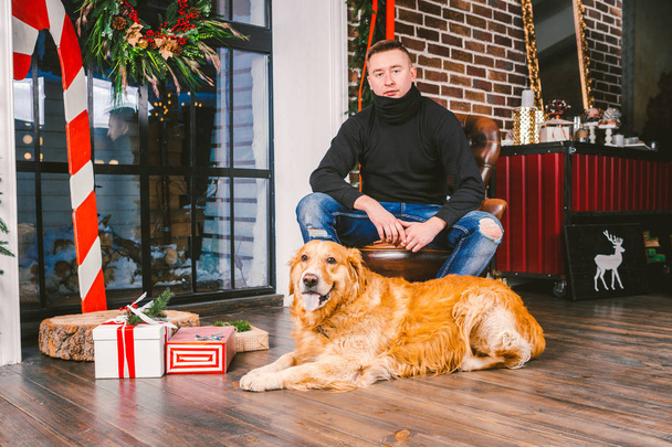 The theme is the friendship of man and animal. Caucasian young male and pet dog breed Labrador Golden Retriever at home inside in winter. Christmas time, New Year holidays. - Foto, afbeelding