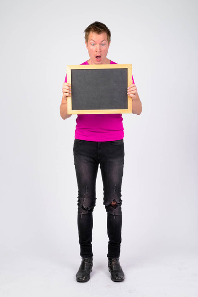 Full body shot of young handsome man looking shocked while showing blackboard - Fotoğraf, Görsel