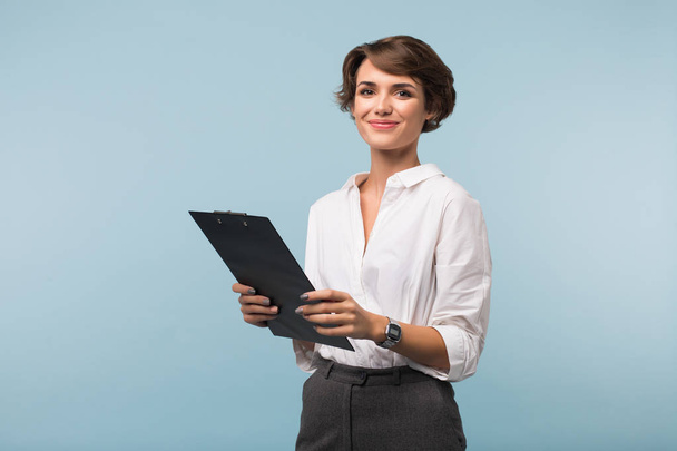 Beautiful business woman with dark short hair in white shirt holding black folder in hands joyfully looking in camera over blue background - Photo, Image