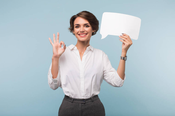 Young cheerful woman with dark short hair in shirt holding white paper shape of message in hand near head happily showing ok gesture on camera over blue background - Valokuva, kuva