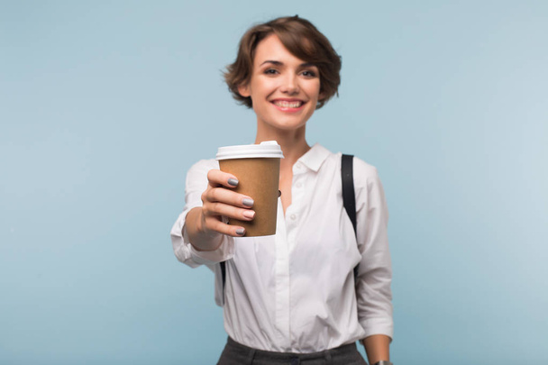 Chica sonriente con el pelo corto oscuro en camisa blanca mostrando felizmente en la cámara taza de café para ir sobre fondo azul
 - Foto, imagen